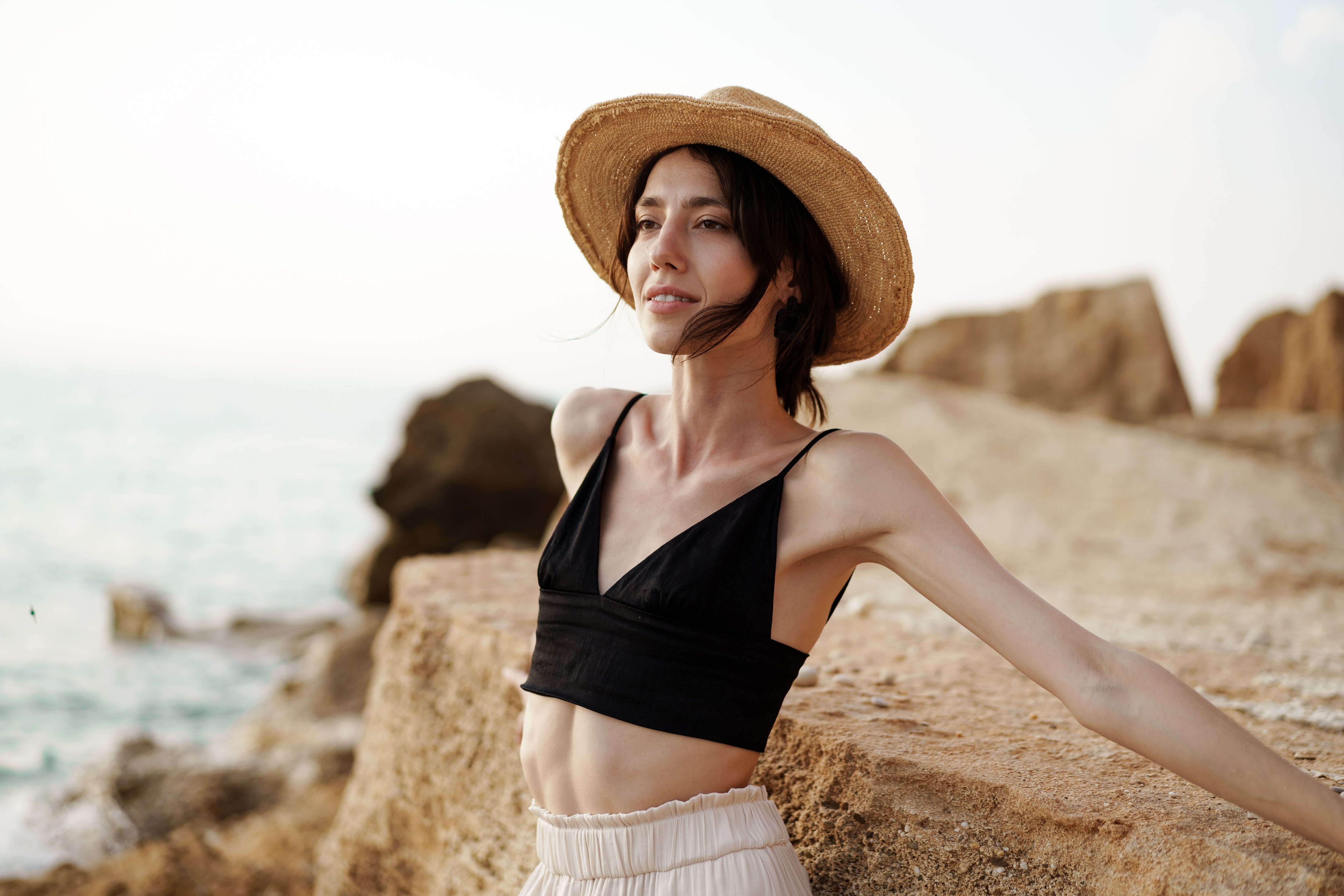 Woman in a straw hat and black bralette leaning on rocks by the sea.jpeg
